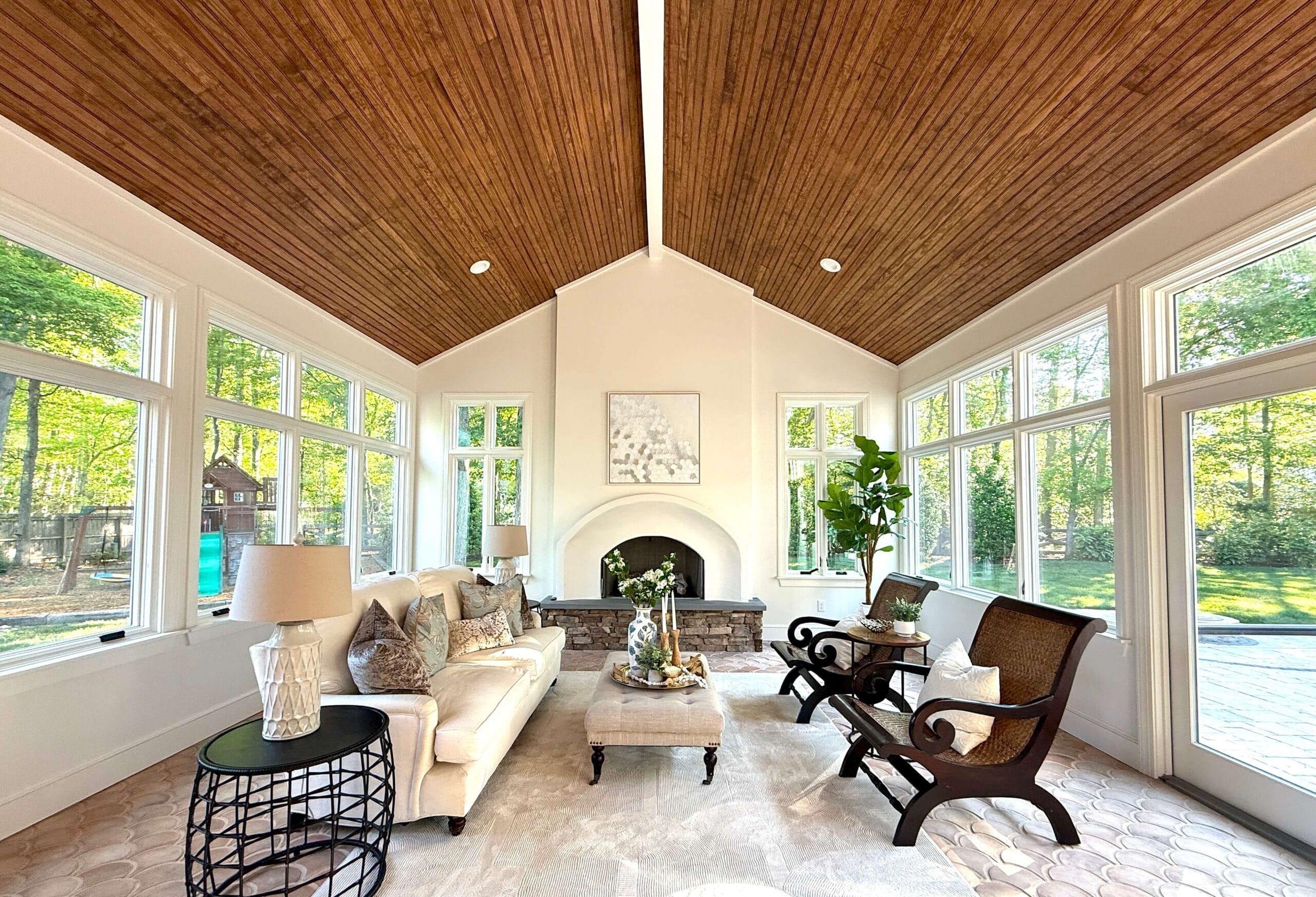 Sunroom with dark wood ceiling, lots of windows, a fireplace, white sofa, coffee table, 2 chairs and an end table with a lamp